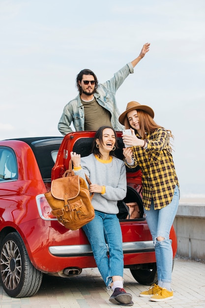 Mujeres que toman autofotos en un teléfono inteligente cerca del maletero del coche y el hombre que se inclina hacia fuera del auto