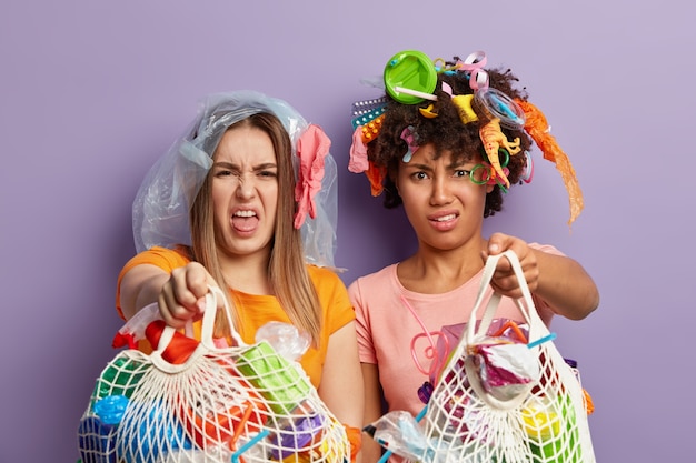 Foto gratuita las mujeres de raza mixta descontentas muestran bolsas de basura con apatía, sienten disgusto, luchan contra el problema ecológico, recogen basura plástica, aisladas sobre una pared purpe. los buenos amigos hacen trabajo voluntario