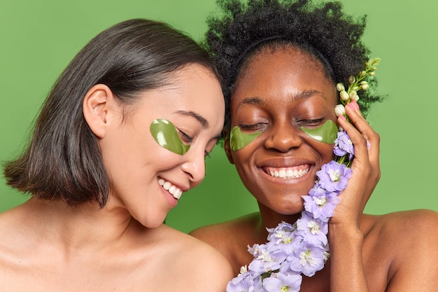 Las mujeres de raza mixta sonríen ampliamente, se aplican parches de hidrogel debajo de los ojos, cuidan la apariencia, sostienen la flor