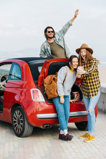 Mujeres riendo tomando selfie en un teléfono inteligente cerca del maletero del coche y el hombre inclinado hacia fuera del auto