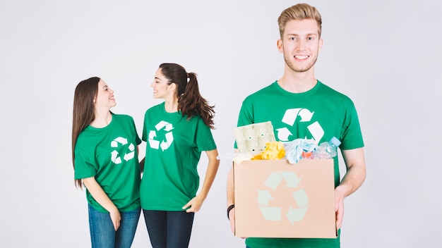 Las mujeres sonrientes detrás del hombre feliz que sostiene la caja de cartón con reciclan artículos