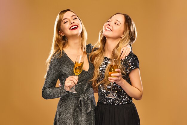 Mujeres sonrientes felices en elegantes vestidos glamorosos con copas de champán en la pared de oro