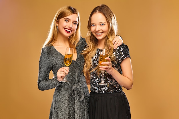 Mujeres sonrientes felices en elegantes vestidos glamorosos con copas de champán en la pared de oro