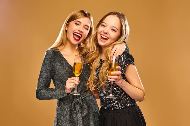Mujeres sonrientes felices en elegantes vestidos glamorosos con copas de champán en la pared de oro