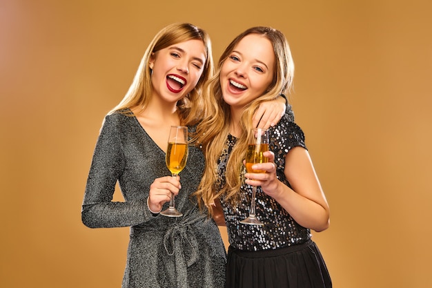 Mujeres sonrientes felices en elegantes vestidos glamorosos con copas de champán en la pared de oro