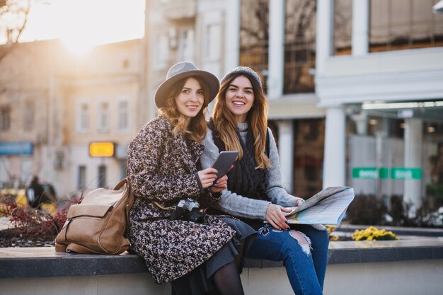 Mujeres sonrientes de moda sentadas en el centro de la ciudad expresando emociones brillantes en un día soleado en la ciudad. Feliz viajando juntos, tratando de encontrar la ubicación en el mapa.