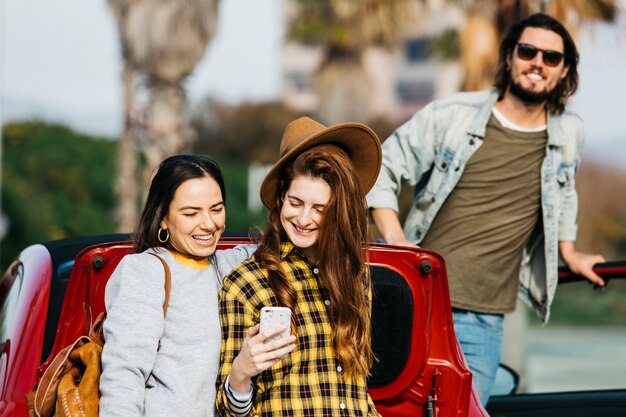 Mujeres sonrientes que toman autofotos en un teléfono inteligente cerca del maletero del coche y el hombre que se inclina hacia fuera del auto