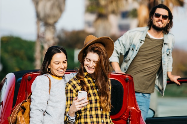 Mujeres sonrientes que toman autofotos en un teléfono inteligente cerca del maletero del coche y el hombre que se inclina hacia fuera del auto