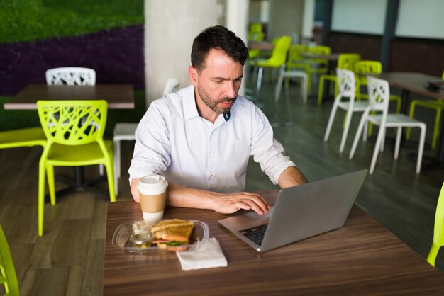 Multitarea para lograr sus objetivos laborales. Ejecutivo de negocios ocupado con ropa informal escribiendo en su computadora portátil mientras se sienta y come una comida en su descanso para almorzar