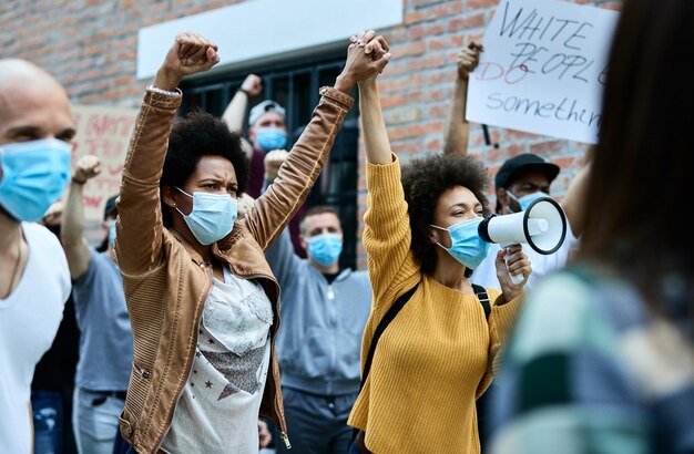 Multitud multiétnica de personas con máscaras protectoras que protestan en las calles de la ciudad El foco está en las mujeres afroamericanas que se toman de la mano