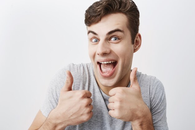 Musculoso hombre caucásico de cabello oscuro positivo en camiseta gris mostrando golpes y sonriendo felizmente con la boca abierta, demostrando sus dientes blancos y parejos. Personas, expresiones faciales y gestos.