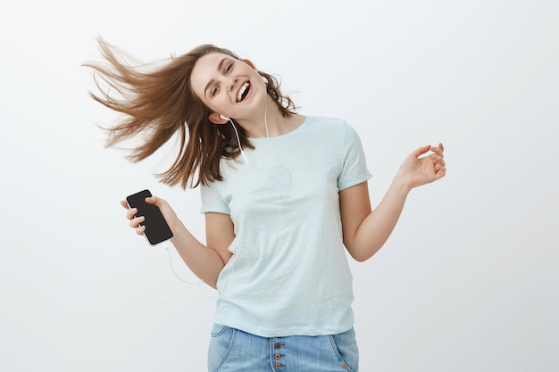 La música es un gran estimulador de emociones. Retrato de encantadora mujer alegre y emotiva saltando alegremente agitando el cabello y sonriendo de alegría escuchando música en auriculares sosteniendo un teléfono inteligente posando contra la pared gris