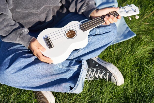 La música y los instrumentos cierran las manos femeninas sosteniendo a un músico de ukelele blanco sentado en el césped al aire libre