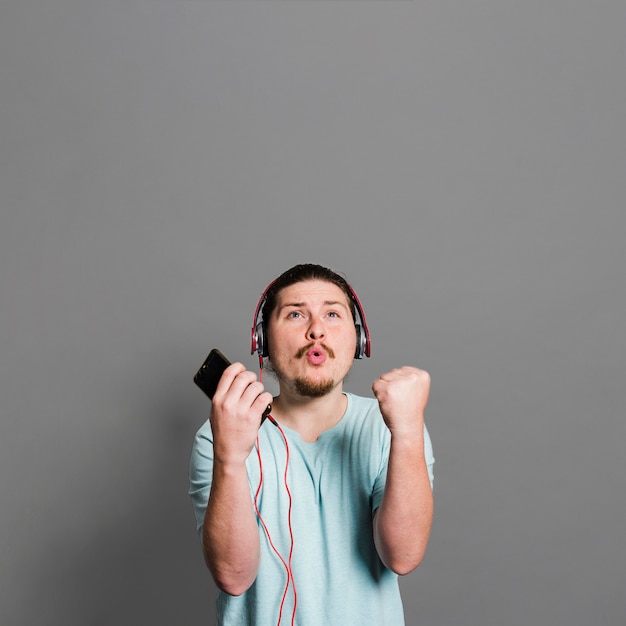 Música que escucha del hombre joven en el auricular que pone mala cara contra la pared gris