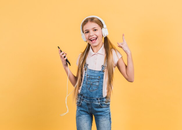 Música que escucha de la muchacha alegre en el auricular que hace la muestra de la roca contra fondo amarillo