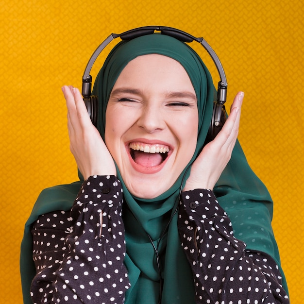 Foto gratuita música que escucha de la mujer árabe joven alegre en el auricular contra fondo amarillo