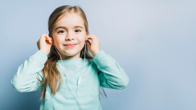 Música que escucha sonriente de la muchacha linda en los auriculares contra fondo azul