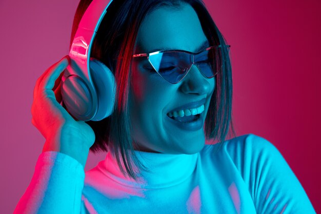 Música. Retrato de mujer caucásica sobre fondo de estudio rosa en luz de neón de moda. Hermosa modelo de mujer con auriculares. Concepto de emociones humanas, expresión facial, ventas, publicidad, moda.