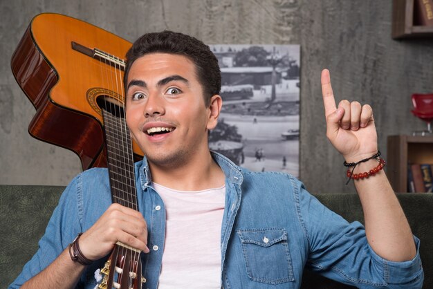 Músico alegre sosteniendo una hermosa guitarra y sentado en el sofá. Foto de alta calidad