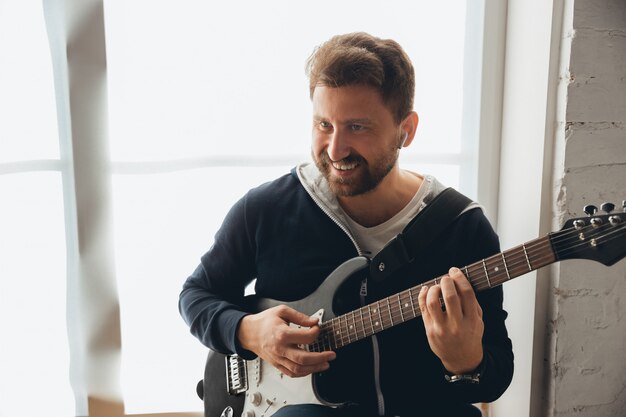 Músico caucásico tocando la guitarra durante un concierto en línea en casa aislada y en cuarentena, alegre improvisación