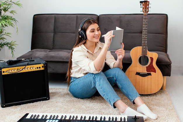 Músico femenino sonriente escribiendo canciones en el bloc de notas junto a la guitarra acústica y el teclado