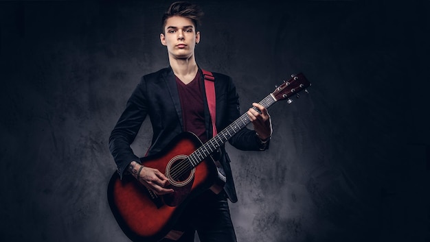 Músico joven con estilo con cabello elegante con ropa elegante con una guitarra en las manos tocando y posando sobre un fondo oscuro.