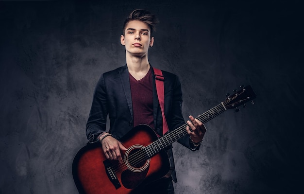 Músico joven con estilo con cabello elegante con ropa elegante con una guitarra en las manos tocando y posando sobre un fondo oscuro.