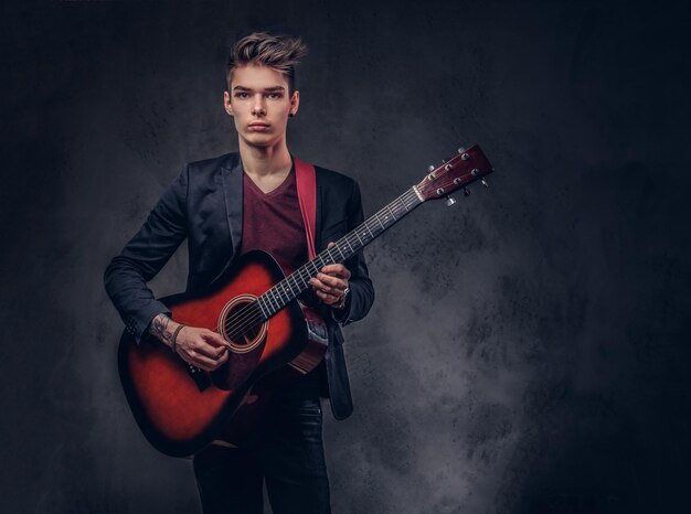 Músico joven con estilo con cabello elegante con ropa elegante con una guitarra en las manos tocando y posando sobre un fondo oscuro.