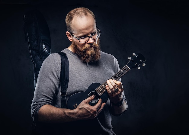 Músico masculino barbudo pelirrojo con gafas vestidas con una camiseta gris tocando un ukelele. Aislado sobre fondo oscuro con textura.