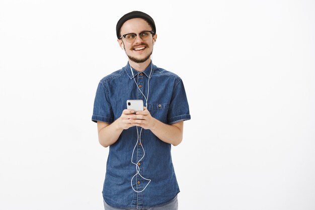 Músico masculino guapo creativo y elegante con gorro negro de moda y gafas escuchando música en auriculares sosteniendo un teléfono inteligente sonriendo curioso y encantado mirando a la derecha nostálgico y complacido