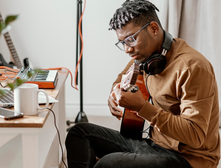 Foto gratuito músico masculino serio en casa tocando la guitarra y mezclando con el portátil