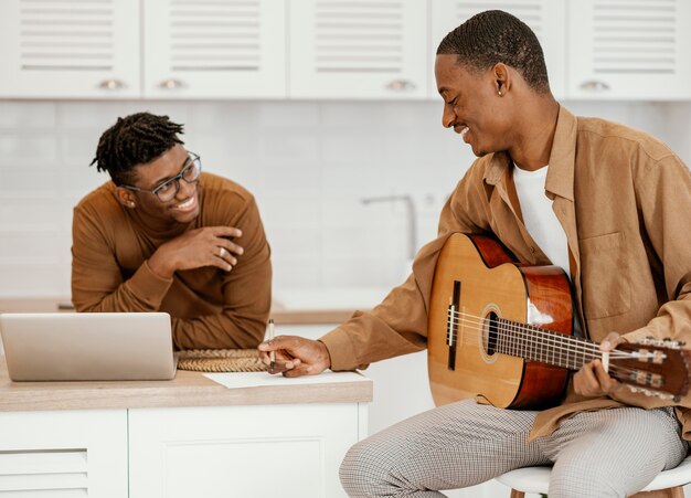 Músico masculino sonriente en casa en silla tocando la guitarra y usando laptop