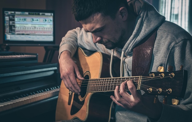 Músico masculino toca la guitarra en casa en el lugar de trabajo cerca de la computadora