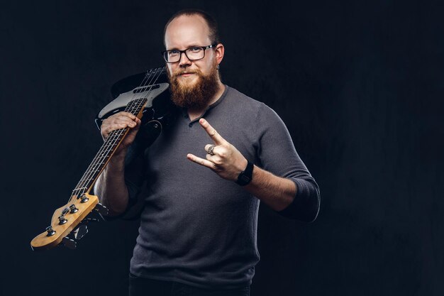 Músico pelirrojo barbudo con anteojos vestido con una camiseta gris sostiene guitarra eléctrica y muestra el signo del rock and roll. Aislado sobre fondo oscuro con textura.