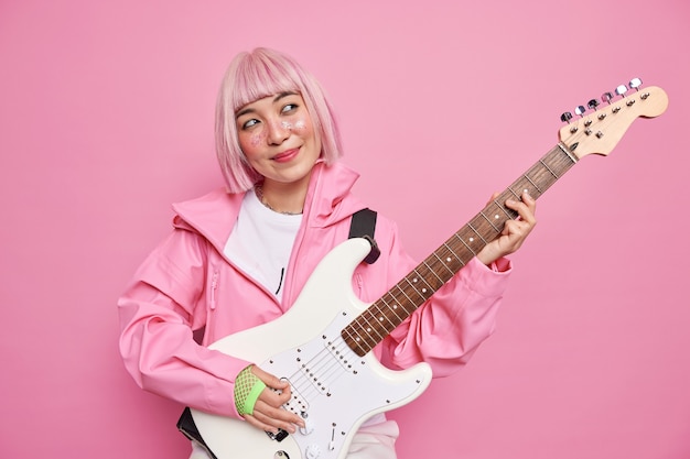 Músico de rock femenino pensativo complacido tocar la guitarra eléctrica blanca realizar una canción popular disfruta de unas vacaciones musicales viste una chaqueta rosa y guantes se encuentra en el interior. artista famoso tiene ensayo antes del concierto