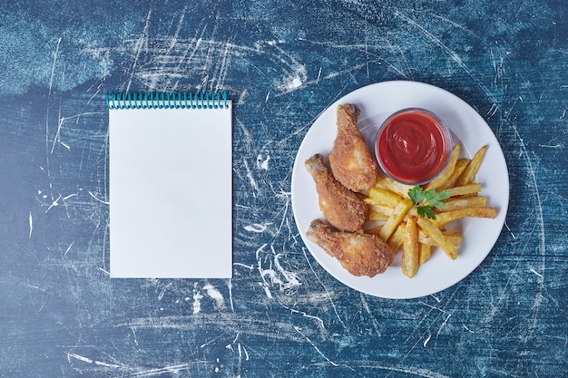 Foto gratuita muslos de pollo y patatas fritas en un plato blanco con un cuaderno a un lado.