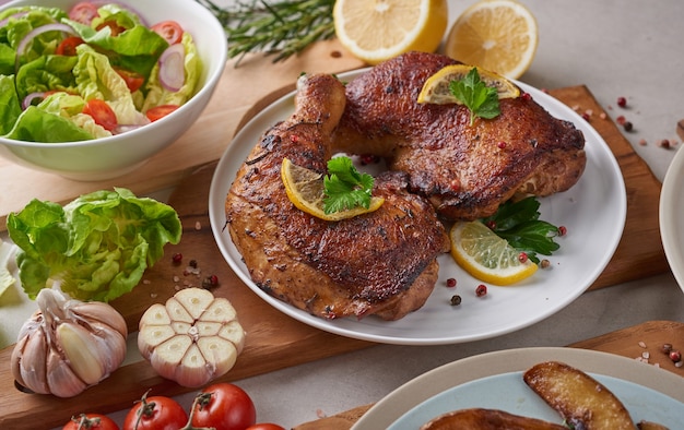 Muslos de pollo a la plancha en salsa barbacoa y verduras asadas y ensalada mixta con tomate, limón en plato blanco sobre mesa de piedra de color claro.