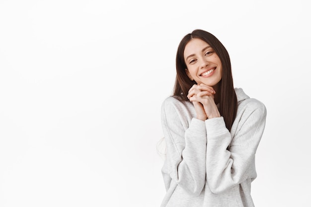 Muy adorable. Linda chica hermosa fascinada por algo, tomados de la mano cerca de la cara y sonriendo, mirando una tierna escena encantadora, viendo un momento romántico, de pie sobre fondo blanco