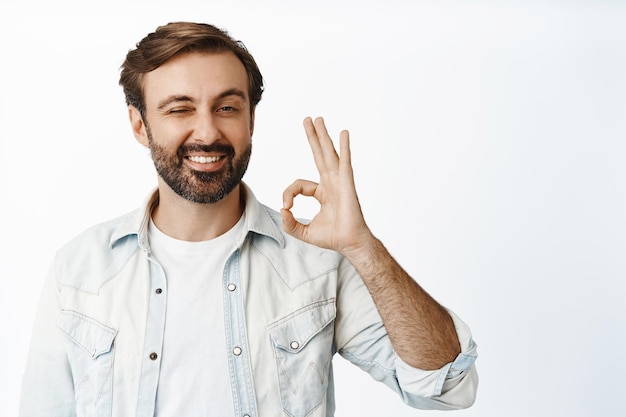 Foto gratuita muy bien un hombre guapo sonriente muestra un signo correcto y guiña un ojo aprueba algo que da permiso recomienda un buen producto fondo blanco