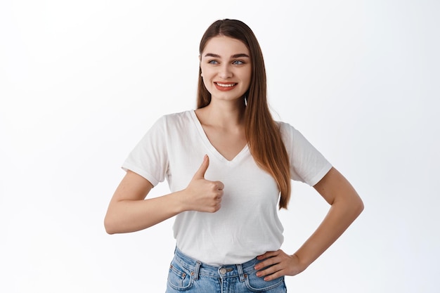 Muy bien. Mujer joven sonriente que mira a un lado la oferta de promoción y muestra los pulgares hacia arriba en señal de aprobación, elogia el buen trato de la tienda, un buen anuncio, satisfecho con su elección, de pie sobre el fondo blanco.