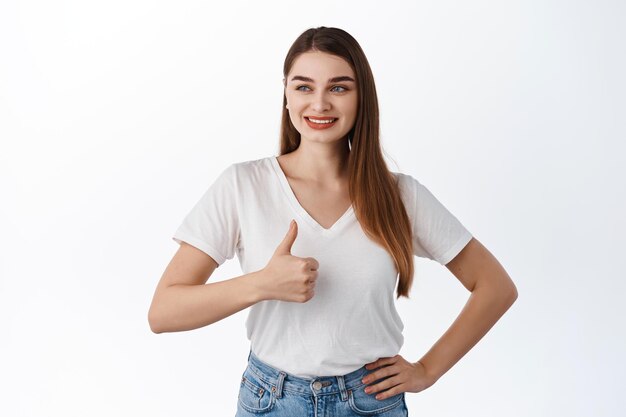 Muy bien. Mujer joven sonriente que mira a un lado la oferta de promoción y muestra los pulgares hacia arriba en señal de aprobación, elogia el buen trato de la tienda, un buen anuncio, satisfecho con su elección, de pie sobre el fondo blanco.