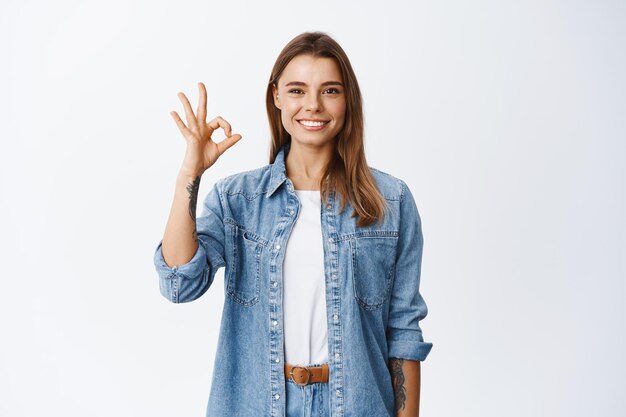 Muy bien. Sonriente joven mujer rubia que muestra un signo bien y parece satisfecho, aprueba lo bueno, de pie satisfecho en blanco, elogia el producto