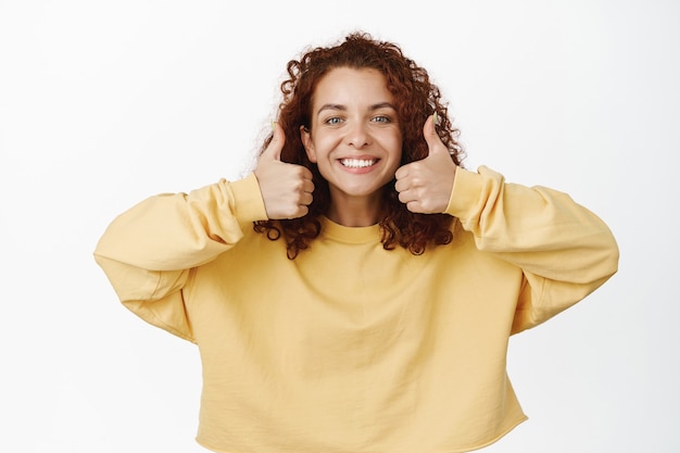 Muy bien. Sonriente mujer satisfecha que muestra los pulgares para arriba cerca de los dientes blancos sonríe, da su aprobación, recomienda algo excelente, de pie sobre blanco