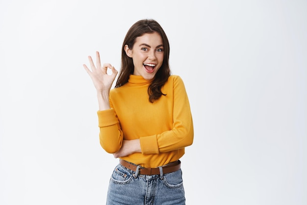 Foto gratuita muy buena modelo femenina joven sonriente con maquillaje natural que muestra un signo de aprobación y dice que sí, confirma que el producto está bien, lo aprueba y le gusta algo sobre fondo blanco