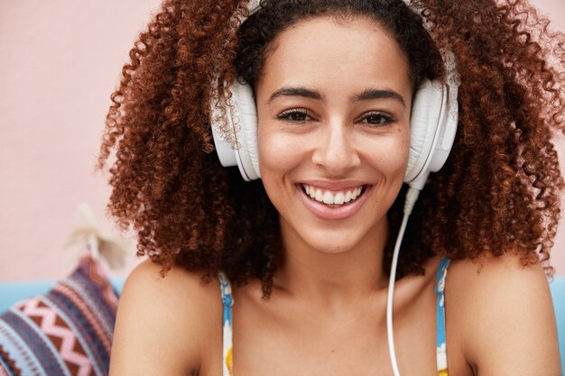 Muy contenta mujer afroamericana con cabello rizado y tupido disfruta de su composición favorita en grandes auriculares modernos