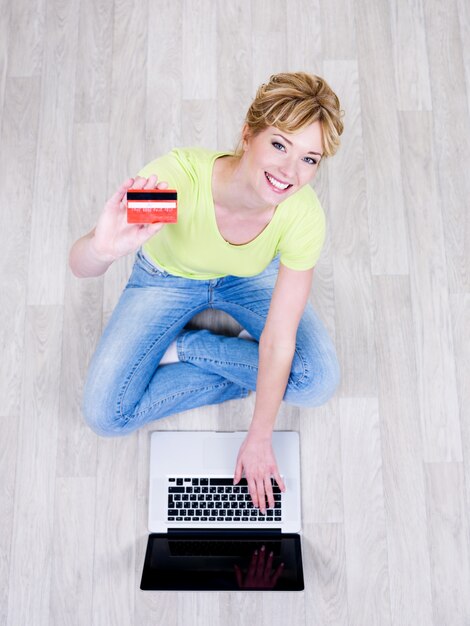 Muy hermosa joven sentada en el suelo con tarjeta de crédito y usando laptop