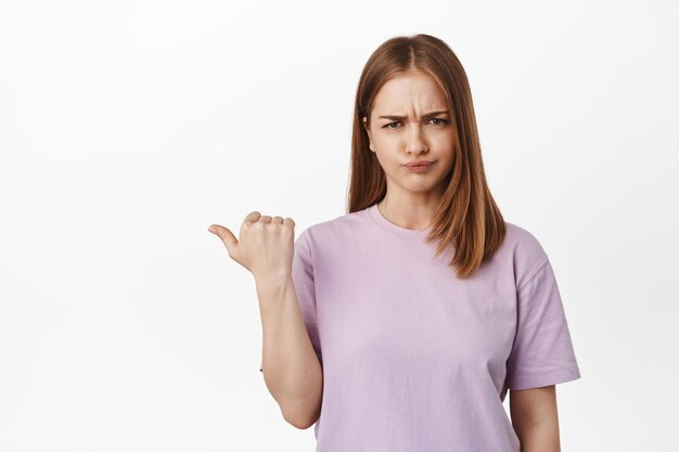 Muy mal. Una chica rubia decepcionada se queja, haciendo muecas y frunciendo el ceño, frunciendo el ceño por disgusto, señalando a la izquierda algo desagradable, de pie disgustada contra el fondo blanco.