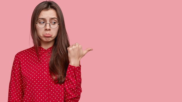 Nada impresionante ahí. Triste mujer ofensiva frunce los labios, apunta a la derecha con el pulgar, vestida elegantemente, se para sobre la pared rosa