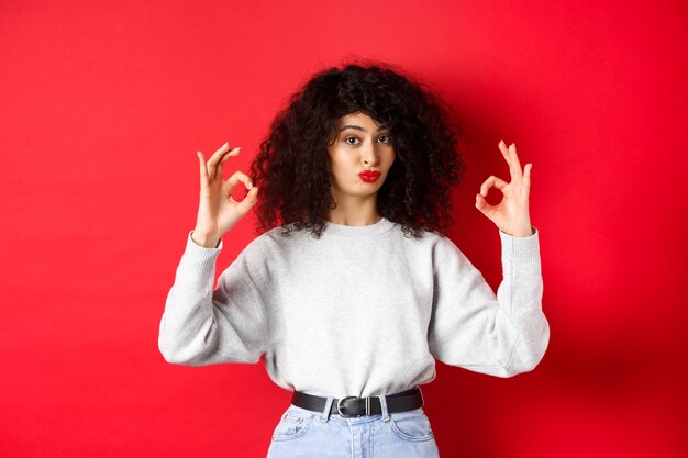 Nada mal. La mujer rizada impresionada asiente con la cabeza en señal de aprobación, frunce los labios y muestra signos de estar bien, como algo bueno, de pie satisfecha sobre un fondo rojo.