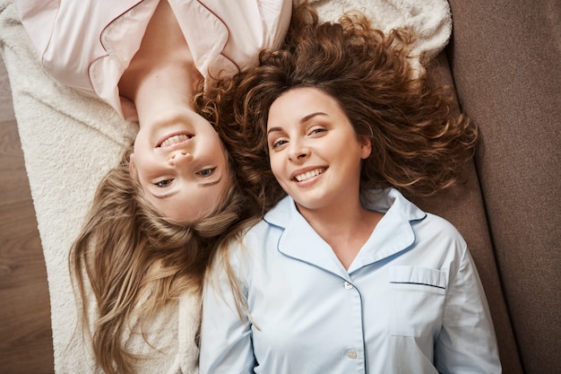 Nada puede ser más fuerte que la amistad. Dos hermosas mujeres europeas tumbadas en el sofá en ropa de dormir cómoda, pasando el tiempo juntas, sonriendo ampliamente, teniendo conversaciones femeninas